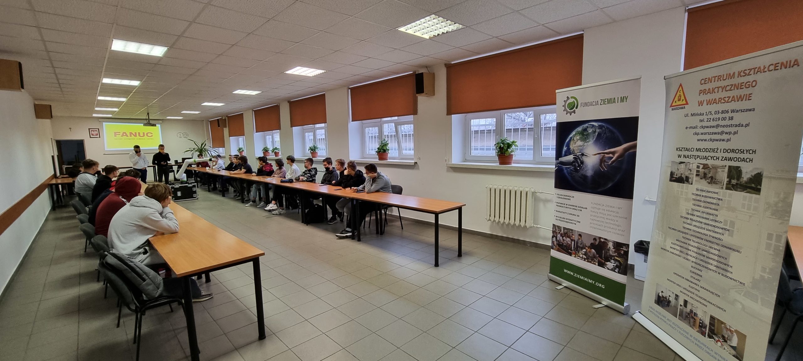 Zdjęcie przedstawia zajęcia dla uczniów szkół ponadpodstawowych w sali szkoleniowej w Centrum Kształcenia Praktycznego w Warszawie wraz z robotem oraz trenerami firmy FANUC.